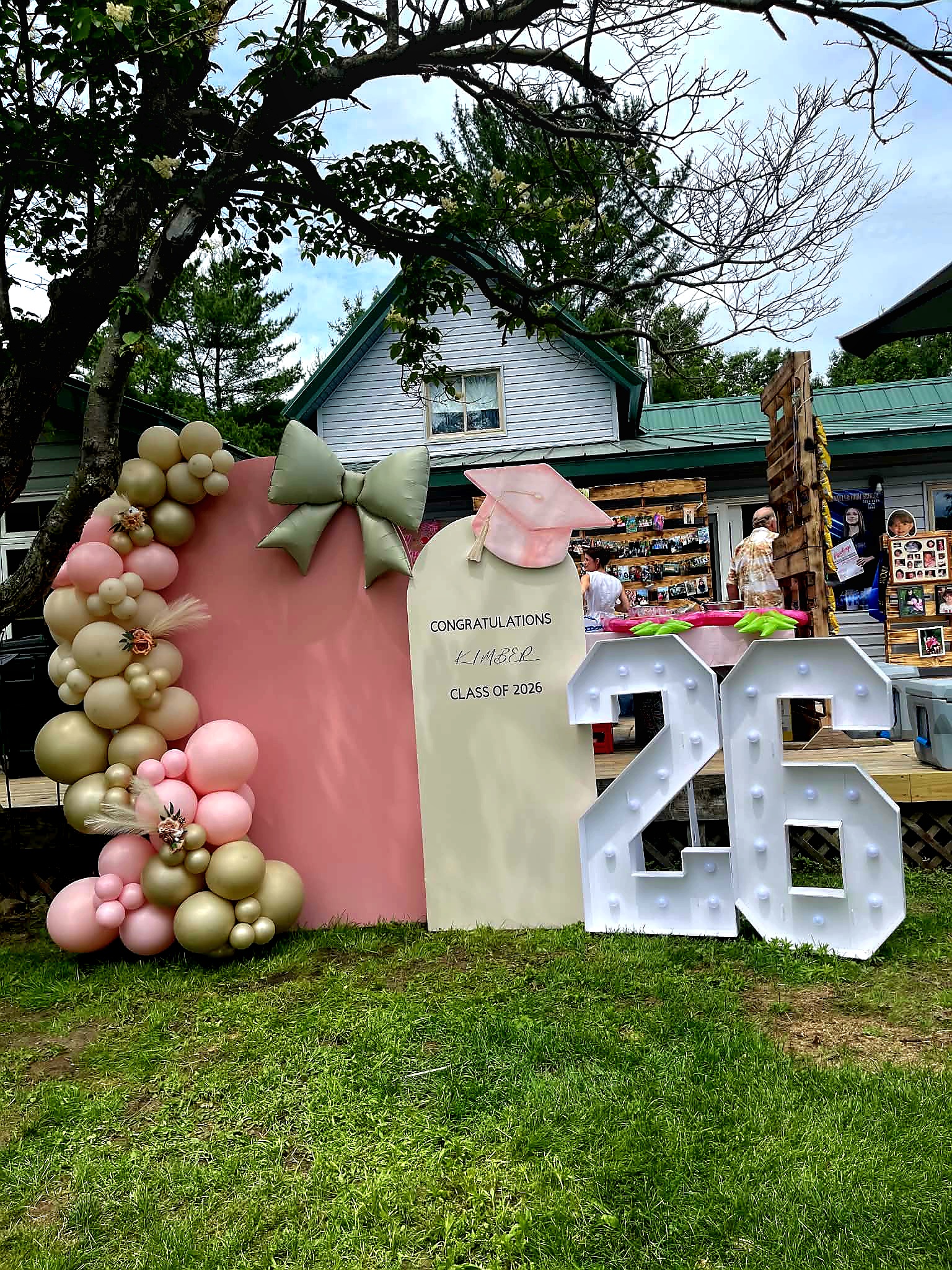 Corrales Designs Double Delight graduation display with soft pink, cream, and gold balloons, marquee 26 numbers, and graduation cap decor